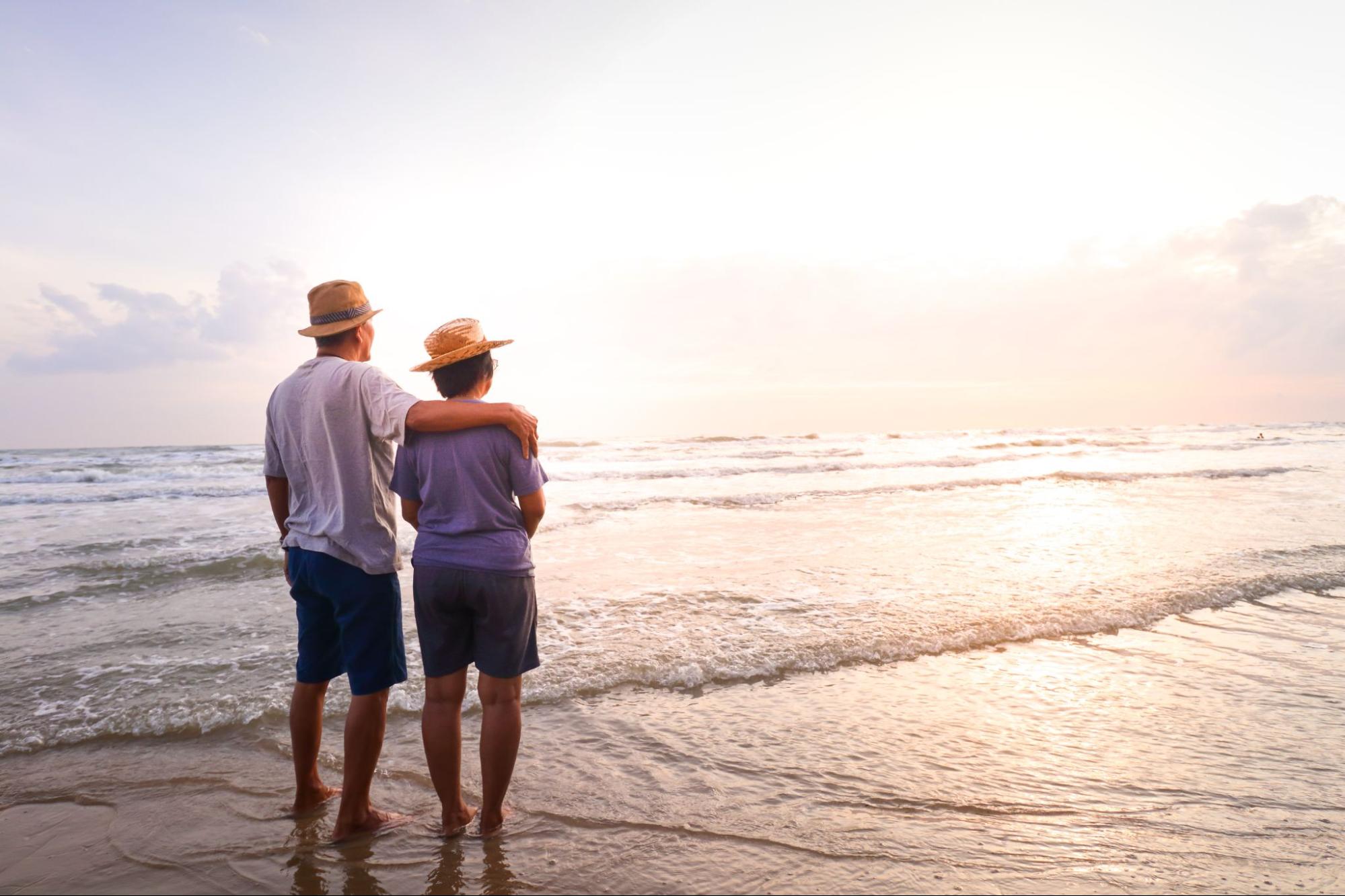 A couple wearing hats standing on the beach and looking at the ocean during sunset.