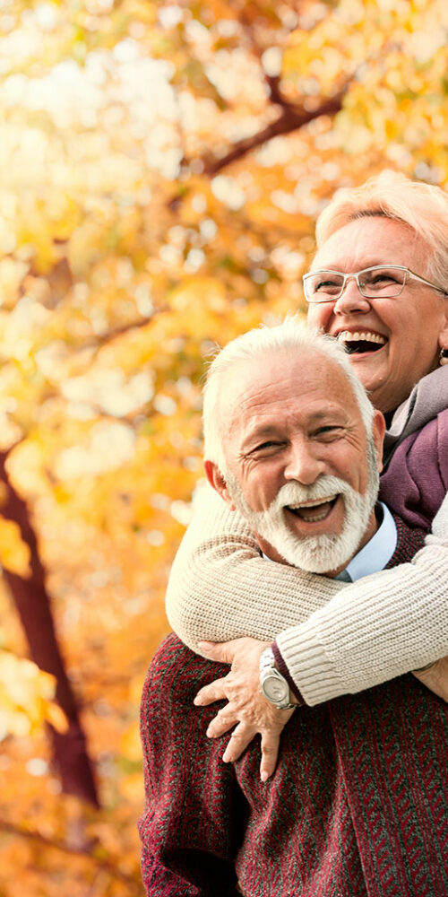 Elderly couple laughing and enjoying a piggyback ride in a park during autumn.