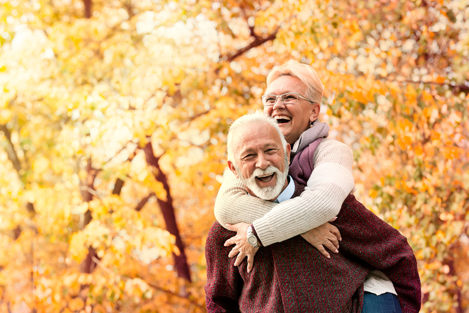 Elderly couple laughing and enjoying a piggyback ride in a park during autumn.