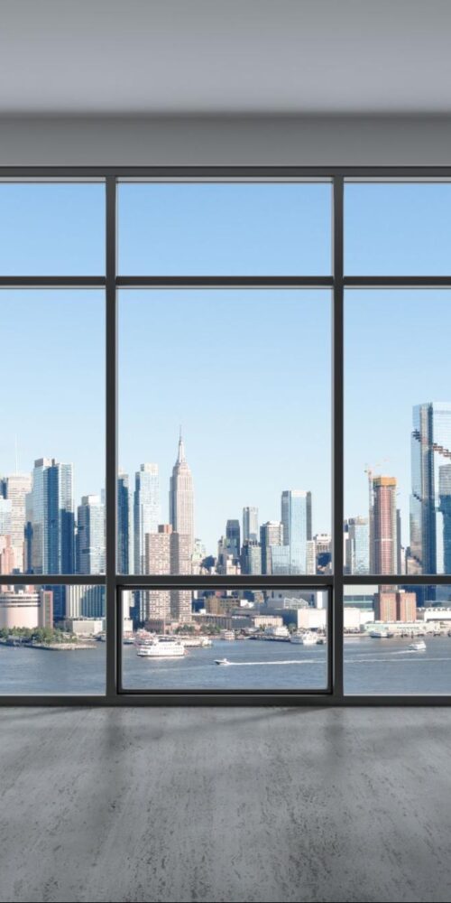 A spacious, modern apartment with large floor-to-ceiling windows offering a panoramic view of the Manhattan skyline and the Hudson River on a clear day.